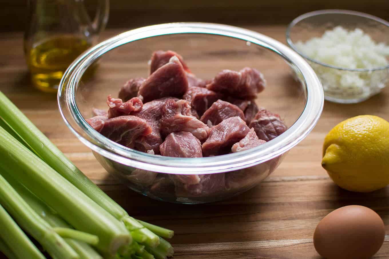 Cubed raw pork shoulder in a glass bowl with celery, olive oil, onions, a lemon, and egg surrounding it.