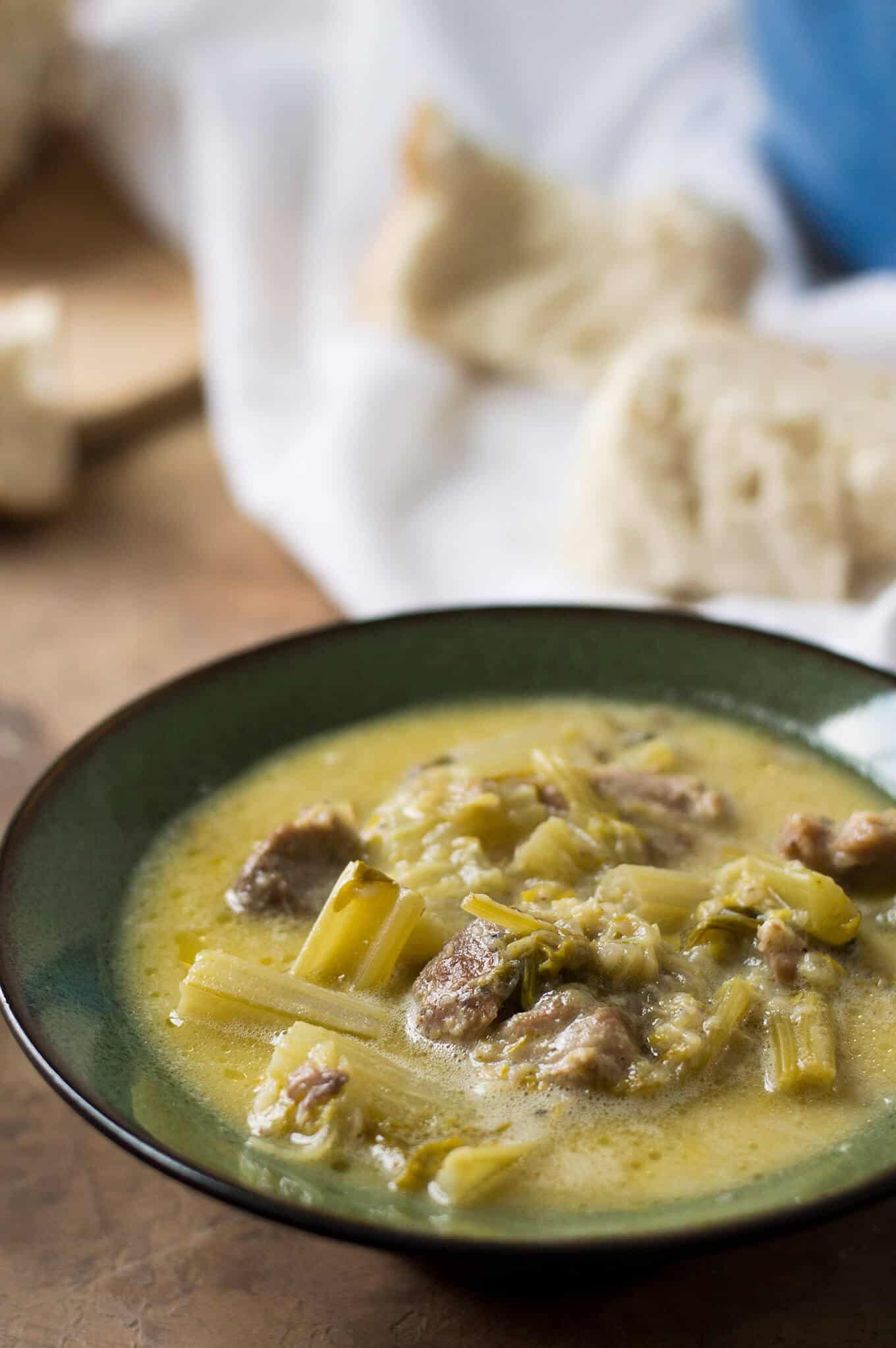 A bowl of pork and celery stew with bread in the background.