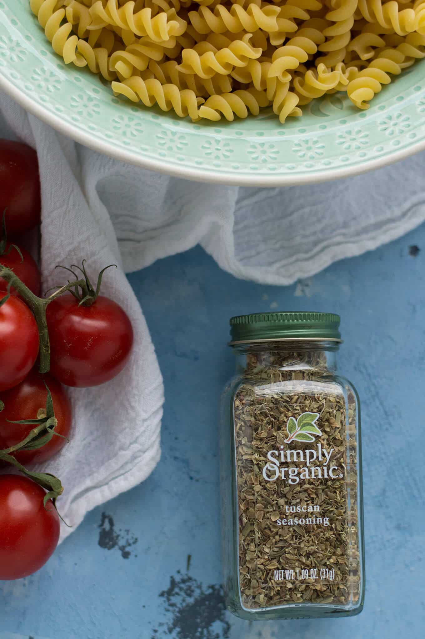 Cherry tomatoes, a bowl of dried pasta, and a bottle of Tuscan seasoning on a table.