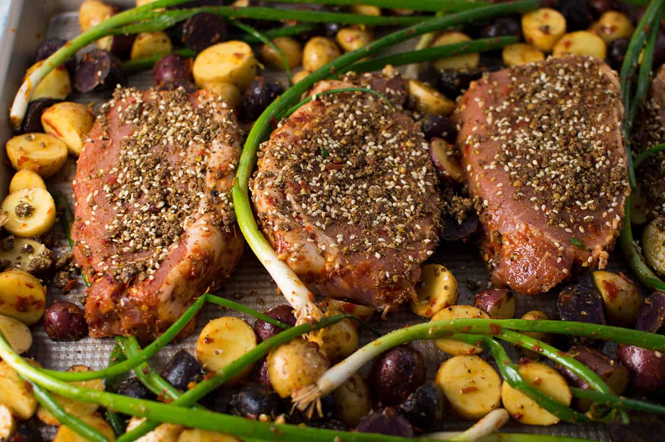 Raw pork chops, scallions, and potatoes on a baking sheet ready for the oven.