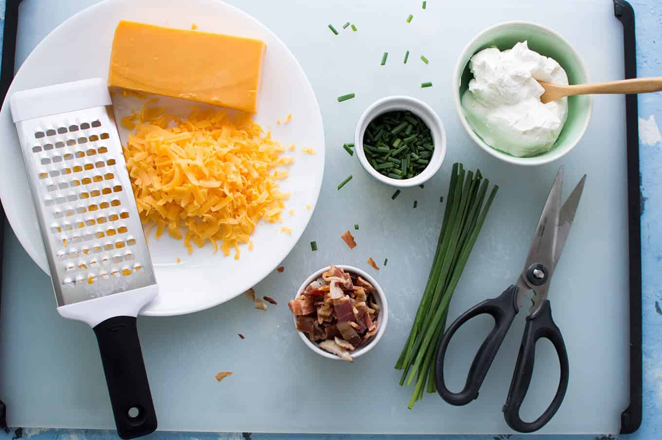 Plates of shredded cheese, chopped bacon, chives, and sour cream on a table.