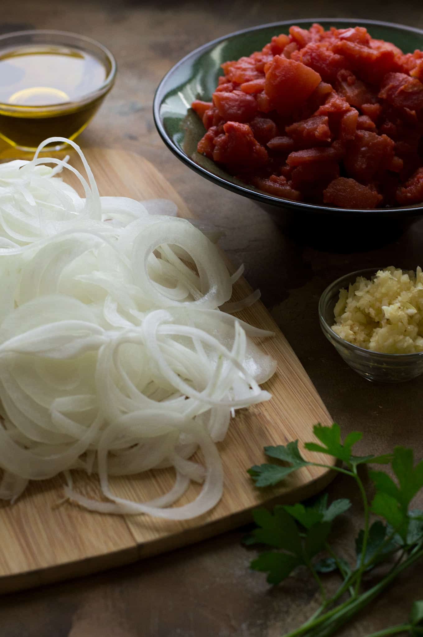 Sliced onions on a cutting board, garlic in a small bowl, and diced tomatoes in a medium bowl.