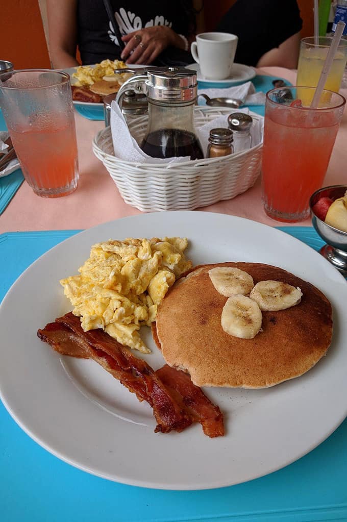 A plate of pancakes, scrambled eggs, and bacon on a table with juice and a basket of syrup, salt, and pepper.