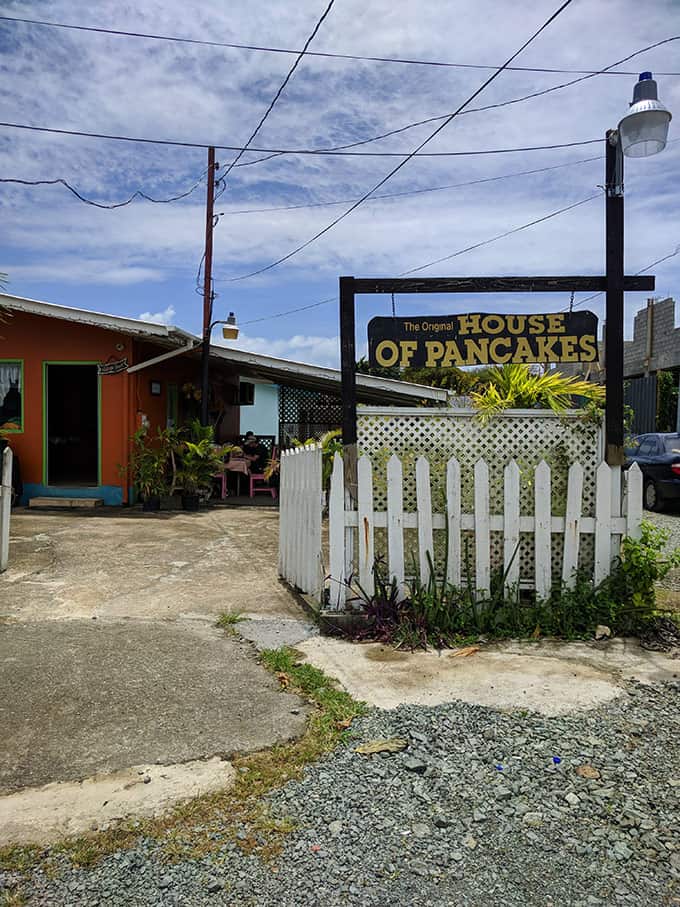 A restaurant sign on a white fence that reads \"The Original House of Pancakes\" with a restaurant in the background.