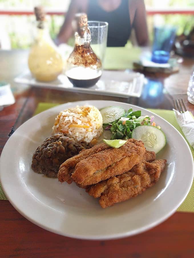 A plate of fried fish, rice, beans, and salad with bottles of hot sauce in the background.