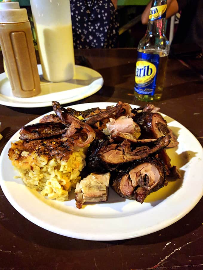 A plate of jerk chicken and macaroni pie on a table with a bottled beer and sauces in the background.