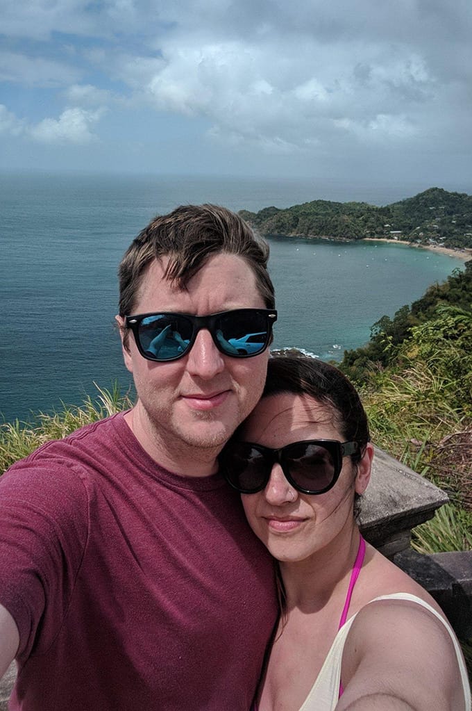 A man and woman wearing sunglasses and standing in front of a cliff overlooking a large body of water.