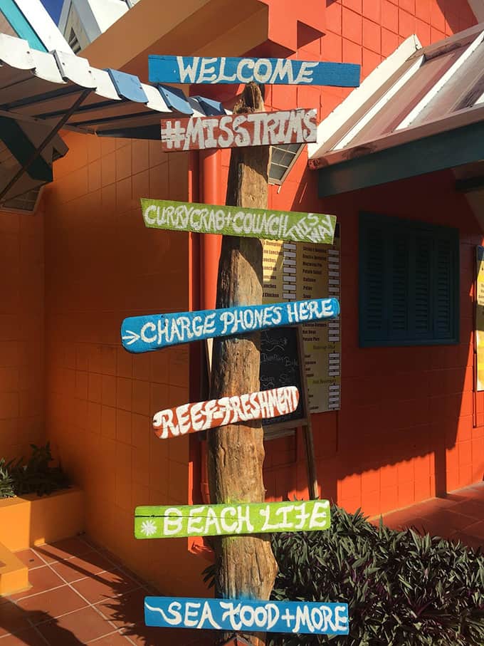 A directional store sign in front of an orange building that reads #misstrims, curry crab + conch this way, charge phones here, refreshment, and beach life.