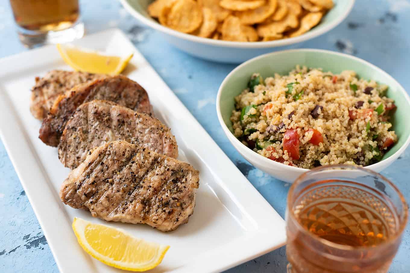 Pork chops, quinoa salad, chips, and cold brew iced tea on a table.