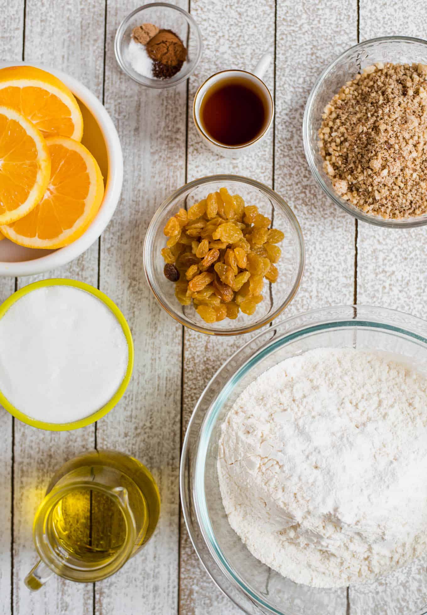 Oranges, sugar, olive oil, golden raisins, ground walnuts, flour, and brandy in various sized bowls for fanouropita.