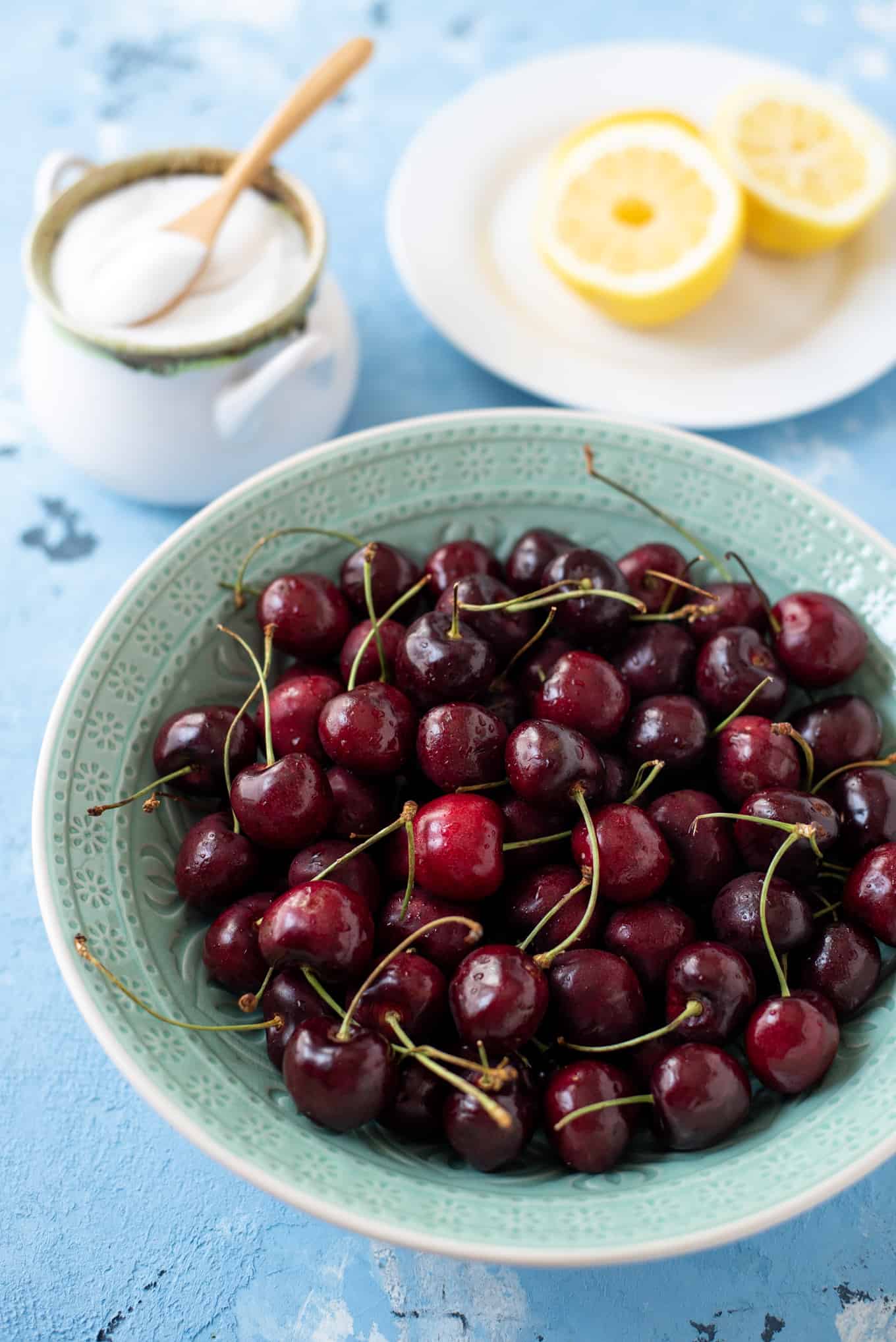 A bowl of cherries on a table with sugar and lemons in the background.