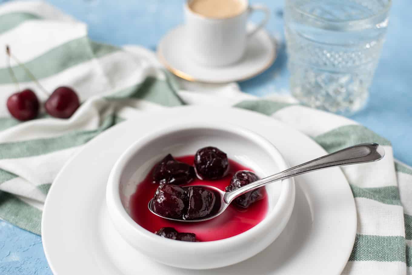 A plate of cherry spoon sweet with a cup of coffee and glass of water on a table.