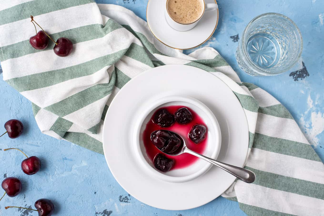 Top-down view of cherry spoon sweet on a plate with a cup of coffee and glass of water on a table.