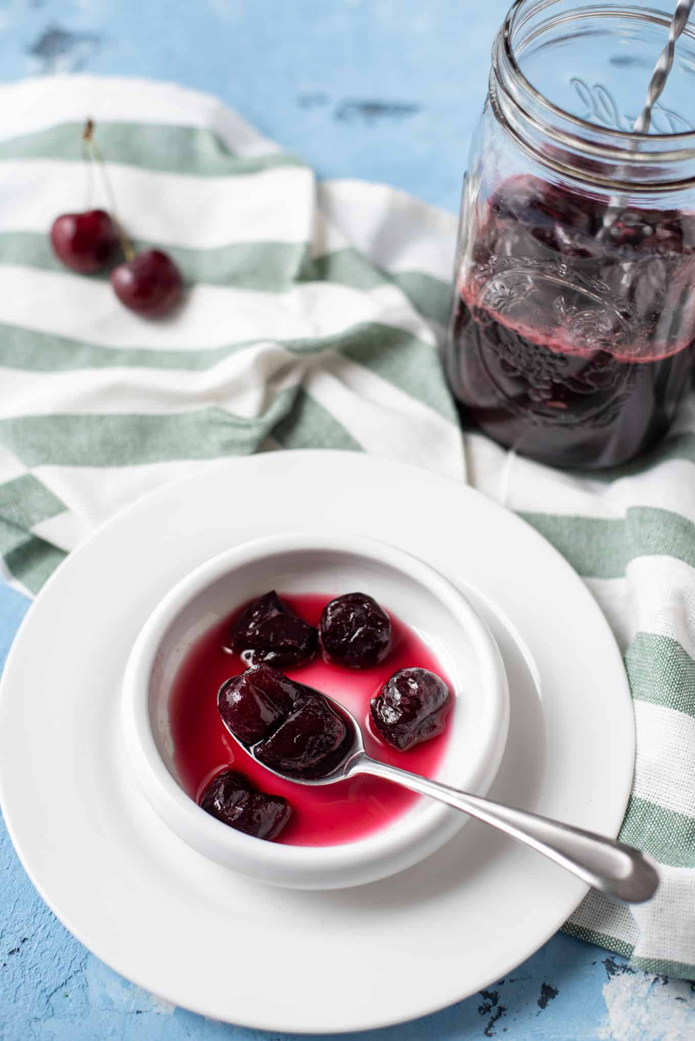 A small white plate with cherry spoon sweet with a jar of preserves in the background.