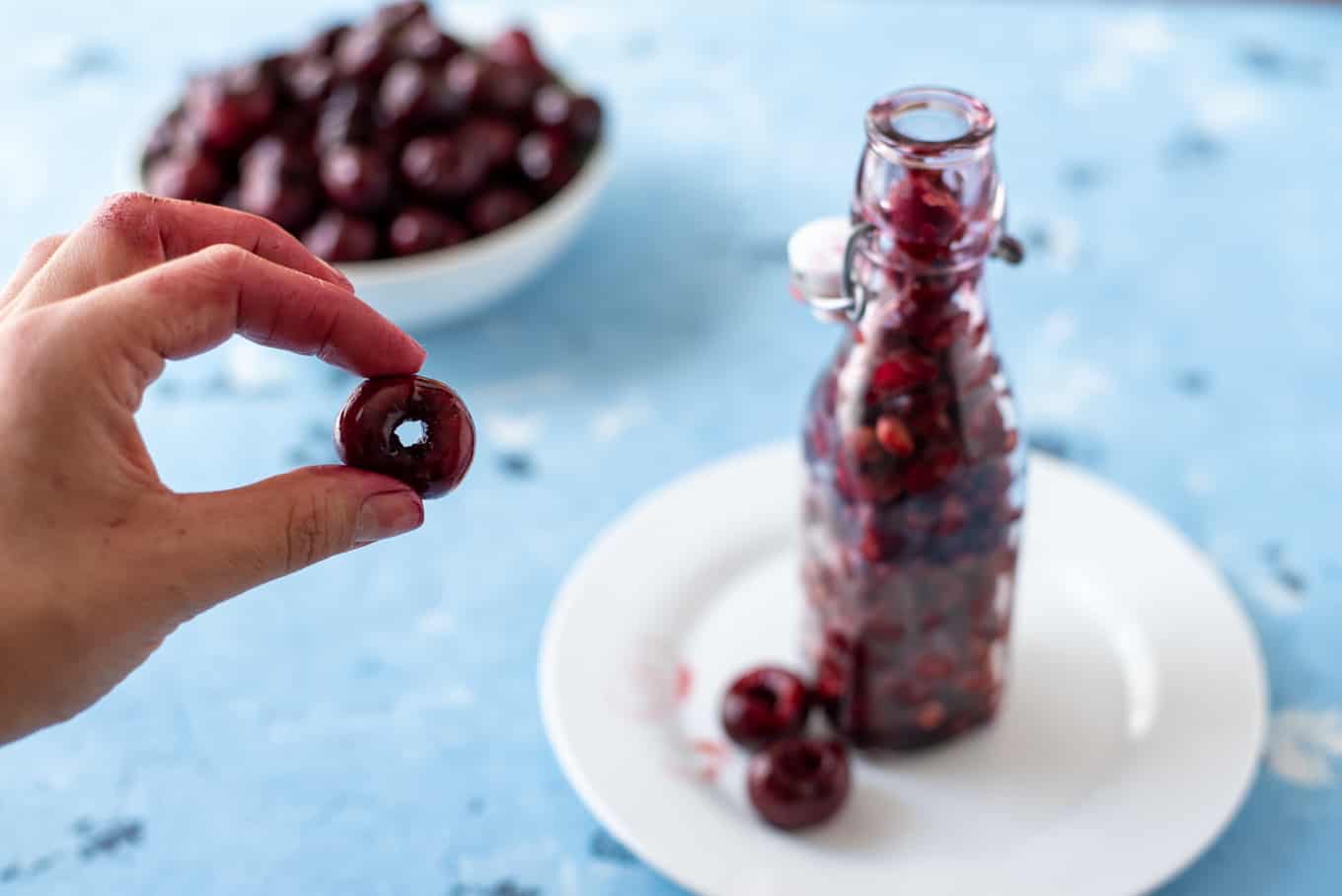 A hand holding a pitted cherry with cherries and pits in the background.