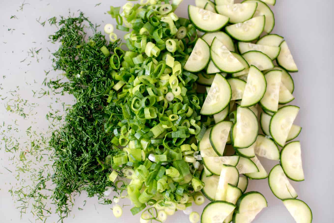 Chopped dill, scallions, and cucumbers on a cutting board.