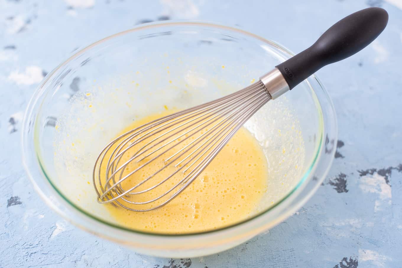 A whisk resting in a glass bowl of mixed eggs.