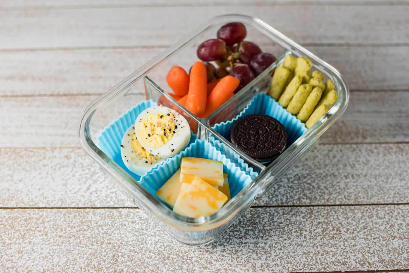 A glass bento box container of cookies, cheese, deviled eggs, fruit, and snacks.