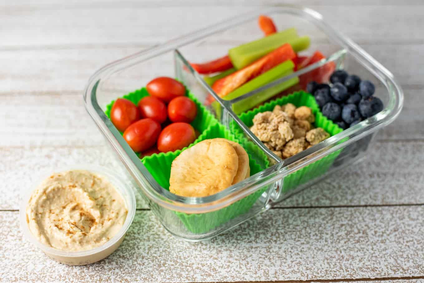 A glass bento box container of crackers, veggie sticks, blueberries, granola, and hummus.