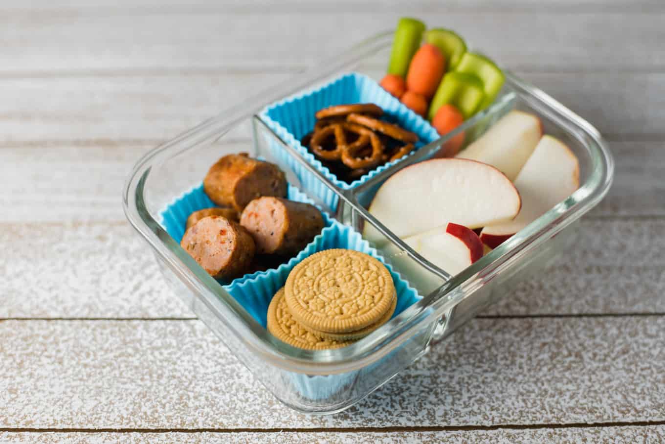 A glass bento box container of cookies, sausage bites, pretzels, apple slices, and veggie sticks.