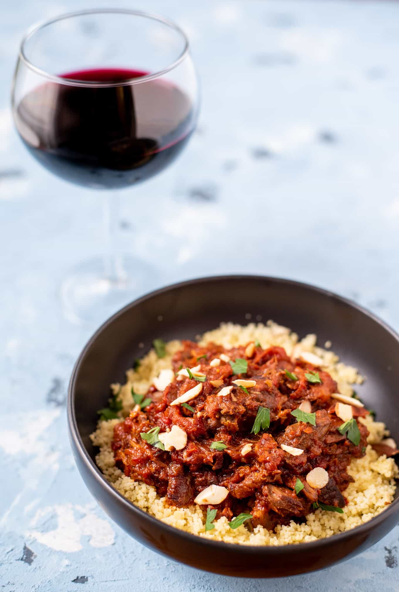 A bowl of lamb tagine with a glass of red wine in the background.