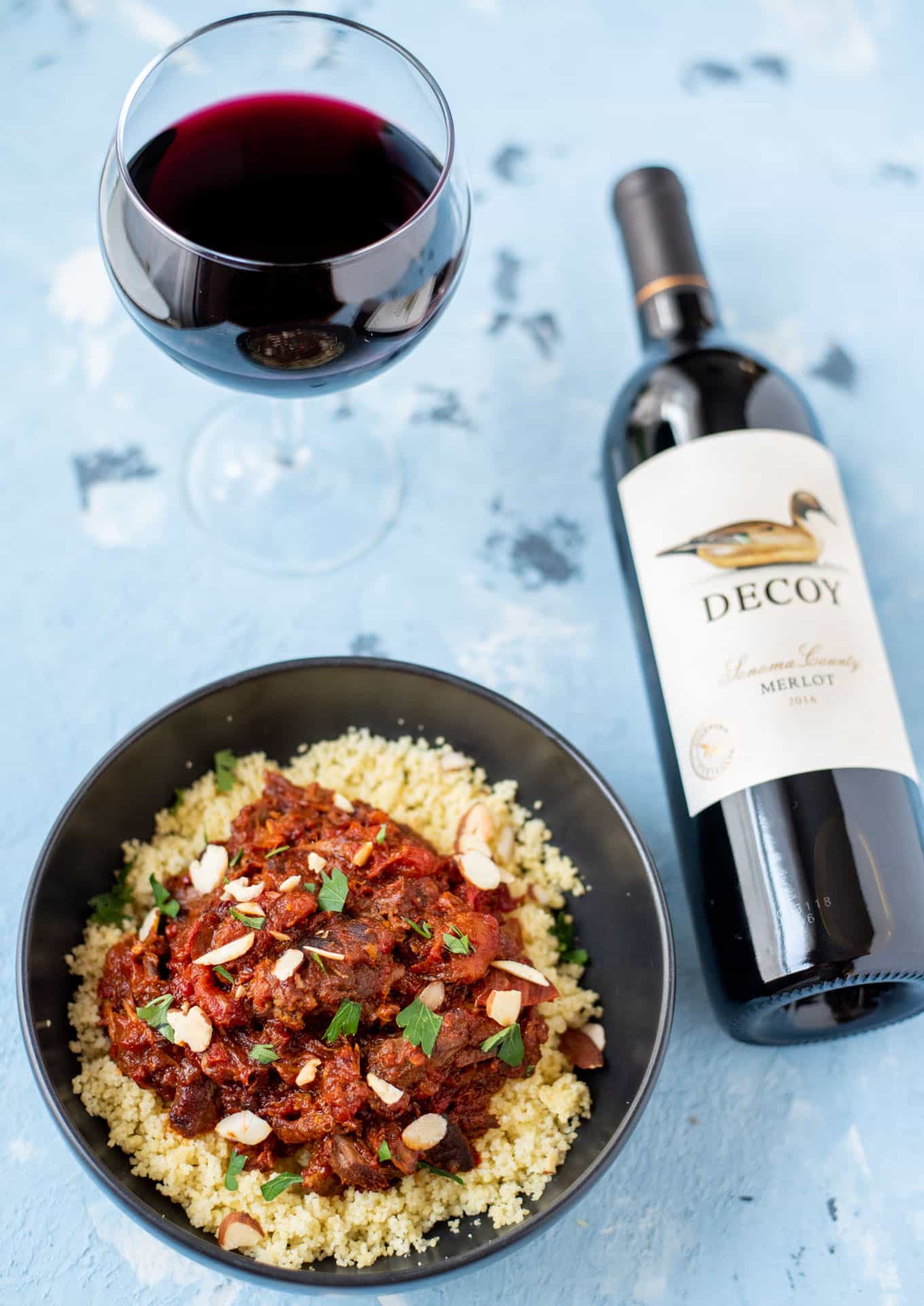 A top-down view of lamb tagine in a bowl, a glass of red wine, and a bottle of red wine.