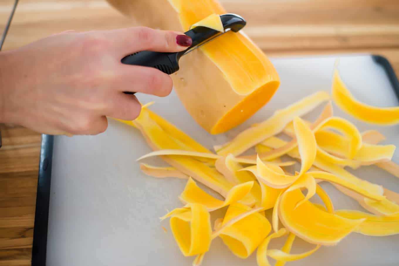 A hand holding a vegetable peeler, peeling a butternut squash on a cutting board.