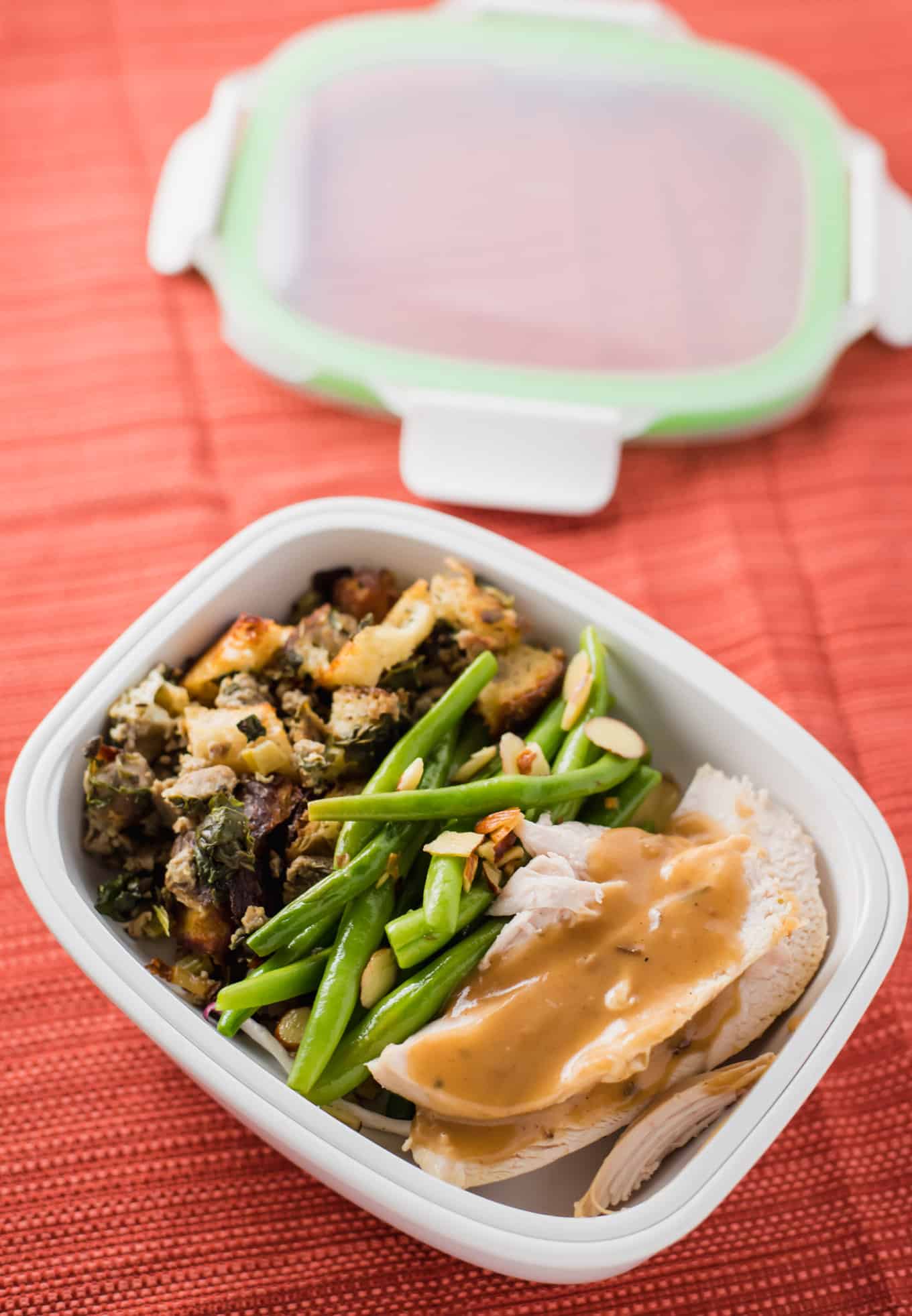 A close-up view of roasted turkey and gravy in a food storage container with green beans and stuffing.