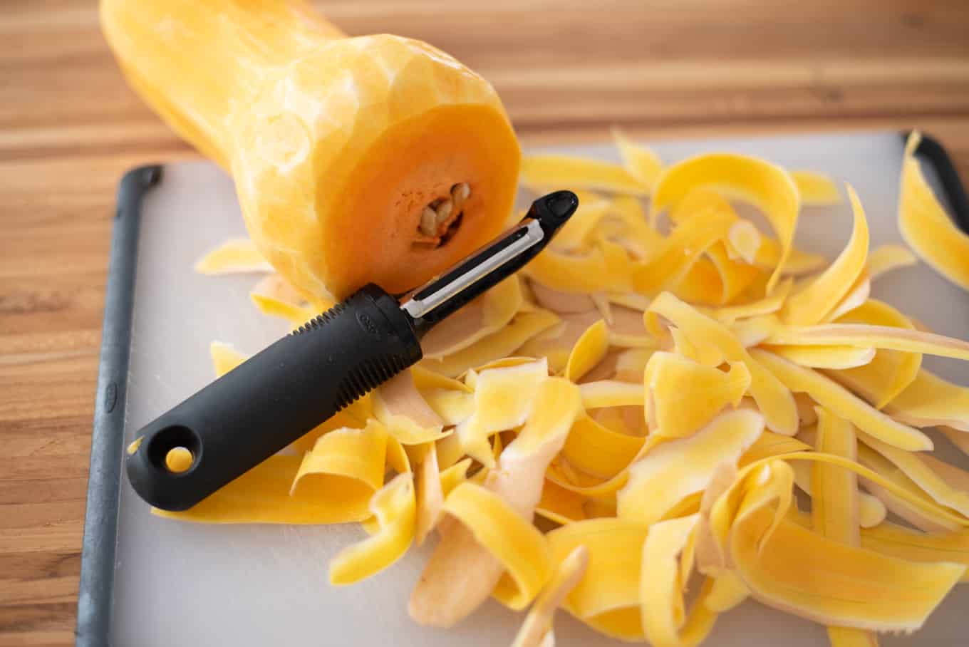 A peeled butternut squash, a vegetable peeler, and peeled scraps on a cutting board.