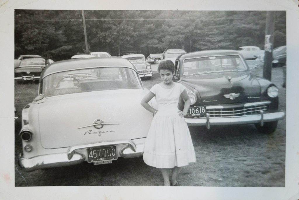 A black and white photo from the 1950s of a woman standing in front of a car.