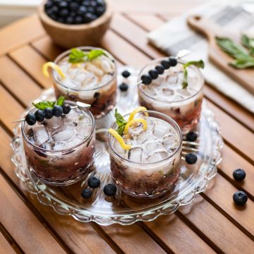 A serving tray with four glasses of blueberry basil bourbon smash. Garnished with fresh blueberries and lemon twist.