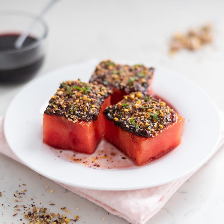A close-up shot of watermelon topped with harissa, dukkah, and mint with pomegranate molasses in the background.