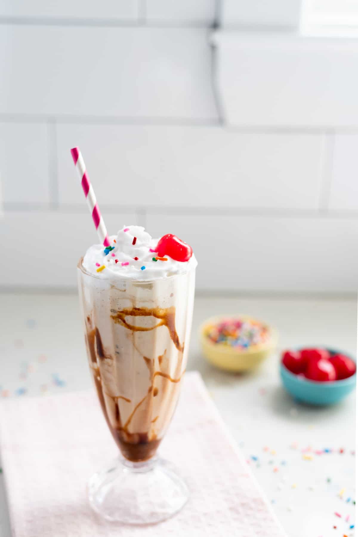 Far-away shot of a vegan vanilla milkshake with a pink and white striped straw. Topped with whipped cream, sprinkles, and a cherry. Extra sprinkles and cherries in the background.