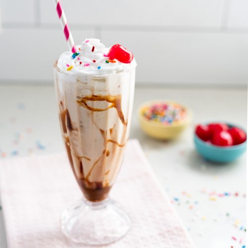 Featured image: A vegan vanilla milkshake topped with whipped cream, sprinkles, and a cherry with extra sprinkles and cherries in the background.