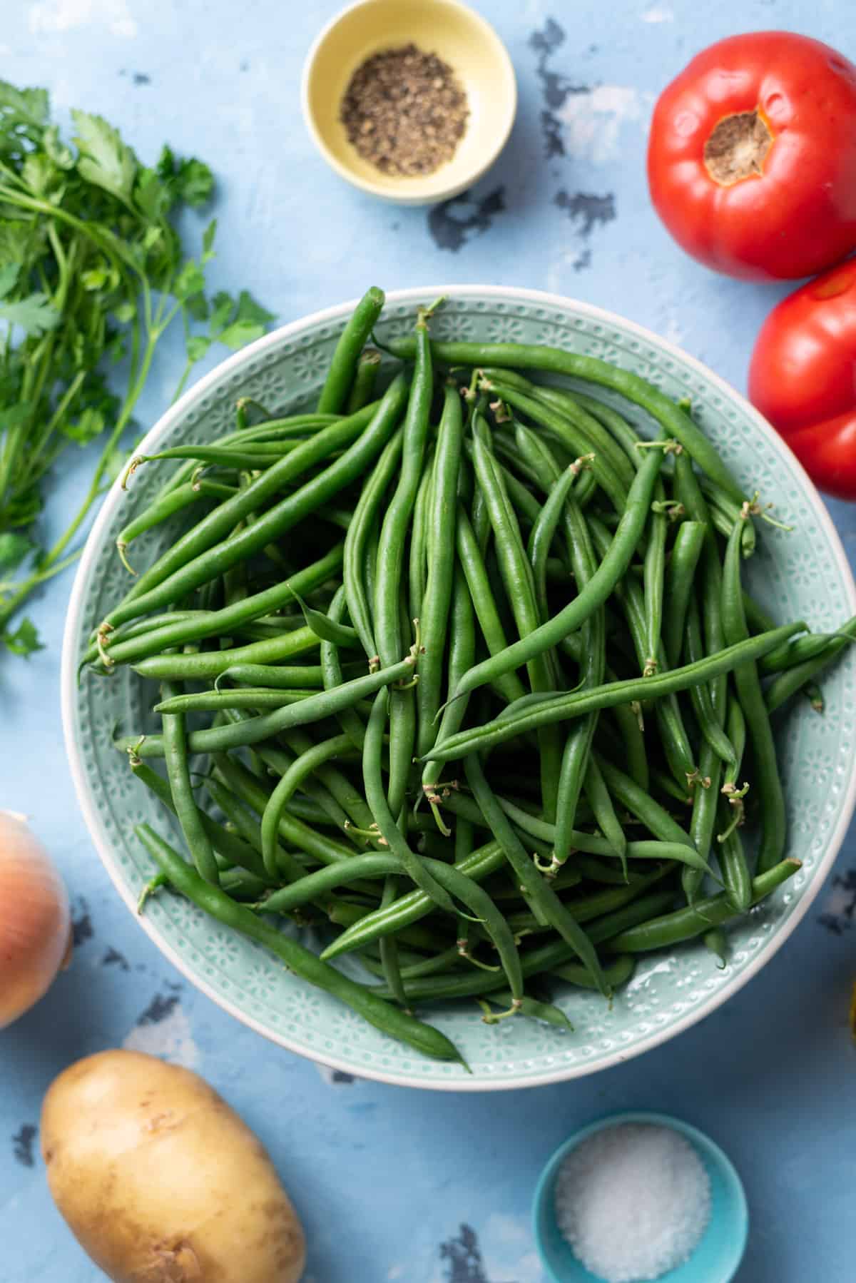 Top-down view of green beans in a blue bowl with tomatoes, potatoes, parsley, and pinch bowls of salt and pepper scattered around.