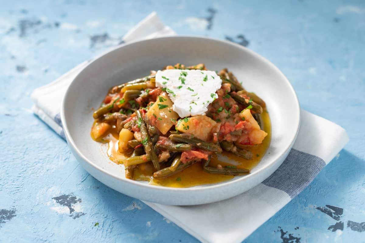 Cooked fasolakia yiahni in a gray bowl topped with feta cheese.