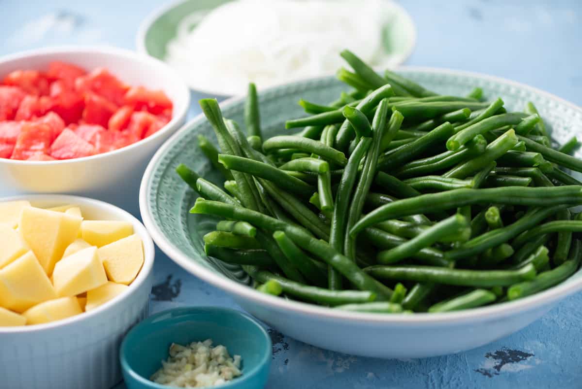 Prepared green beans, cubed potatoes, chopped tomatoes, sliced onions, and minced garlic in prep bowls.