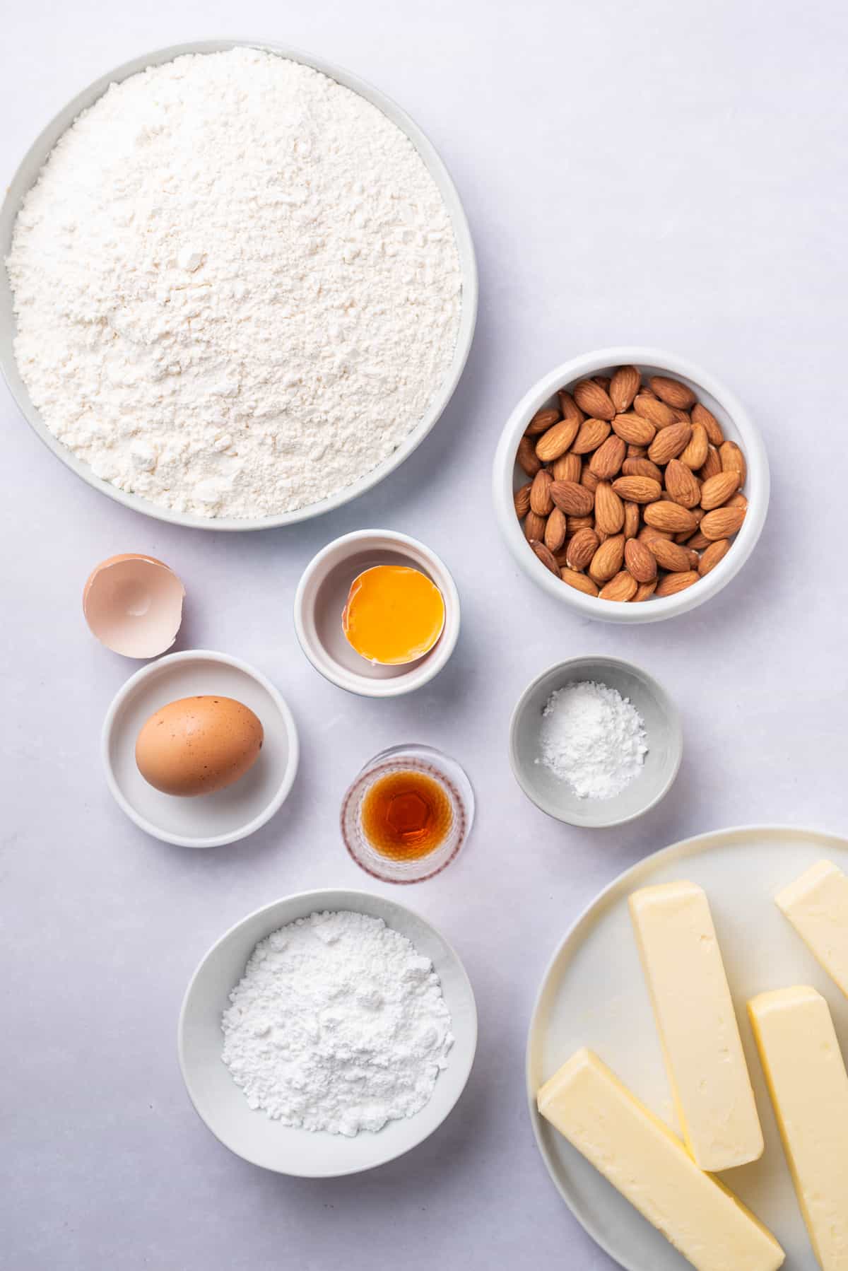 Bowls of flour, eggs, almonds, powdered sugar, brandy, flour, baking soda, and butter on a tabletop.