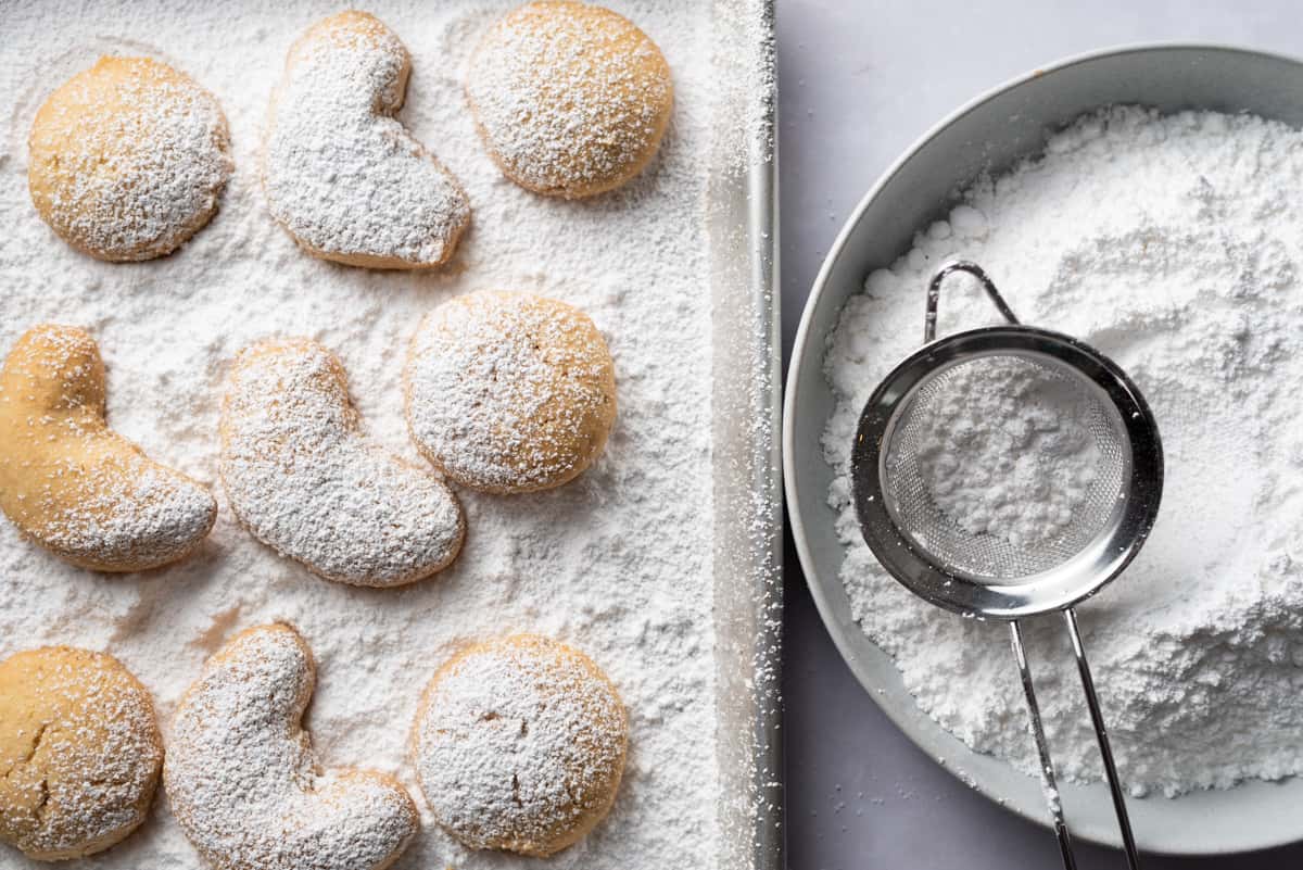 A baking sheet of shortbread cookies partially covered in powdered sugar and a bowl of powdered sugar with a sieve on top.