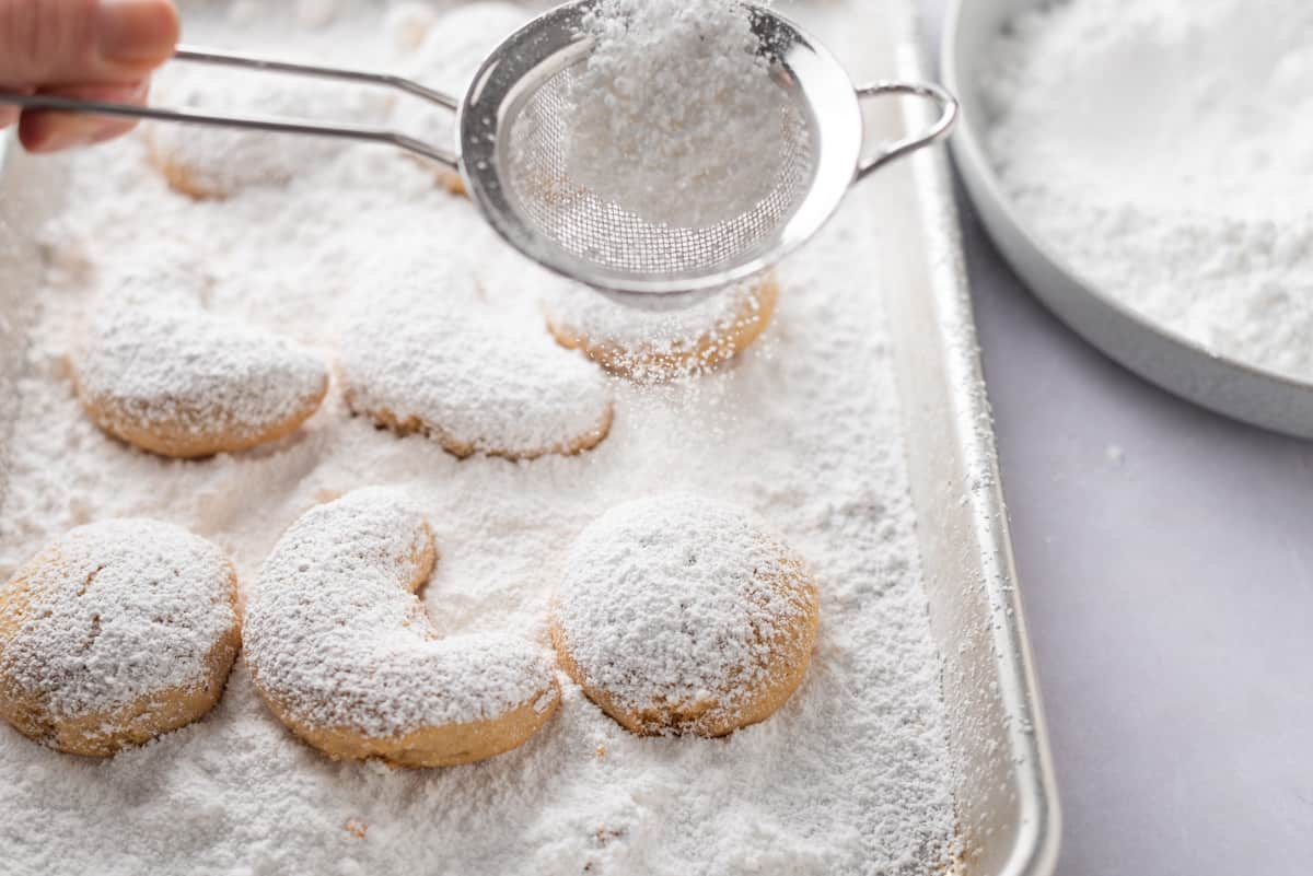 A hand holding a sieve of powdered sugar over shortbread cookies in a baking sheet.