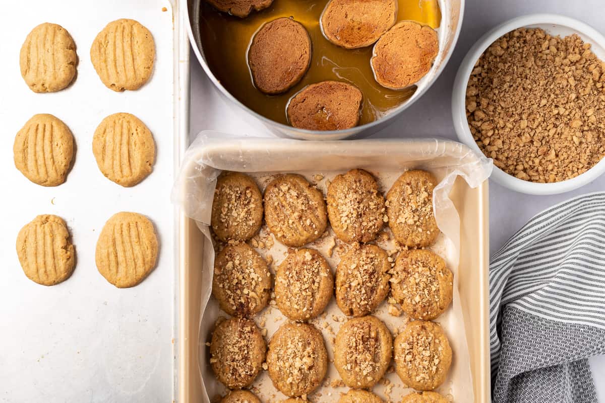 A baking sheet of plain cookies, saucepan of cookies in honey syrup, bowl of chopped walnuts, and wax-lined baking pan of honey-dipped cookies on a tabletop.