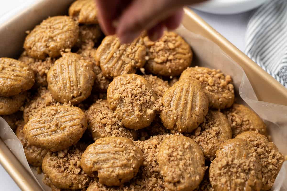 A hand sprinkling chopped walnuts onto Greek honey cookies in a wax-lined baking pan.