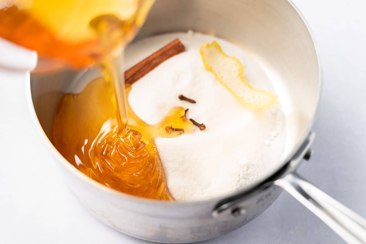 Honey being poured into a saucepan filled with sugar, whole cloves, lemon peel, and a cinnamon stick.