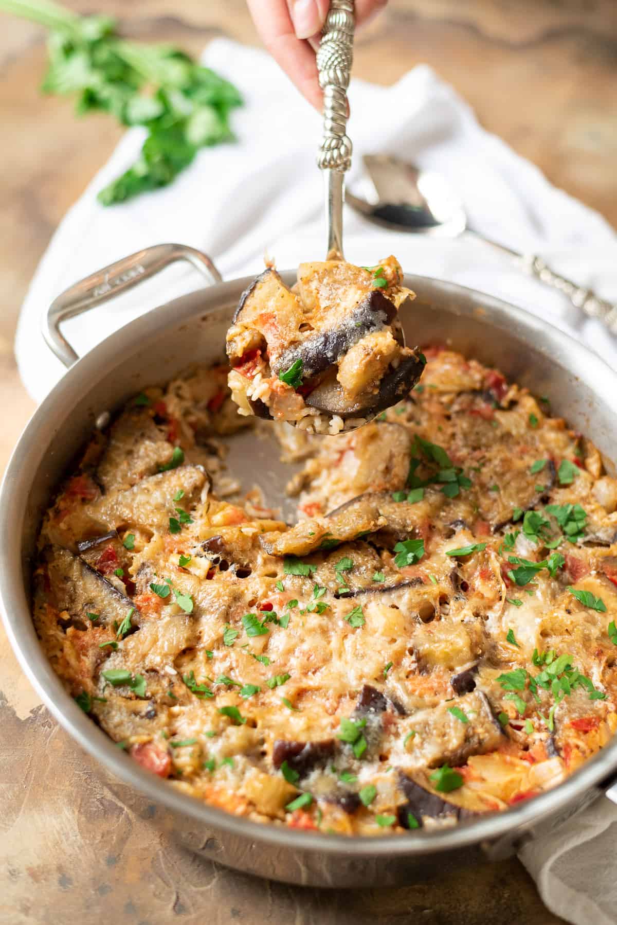 A hand holding a serving spoon with a scoop of baked eggplant and rice casserole.