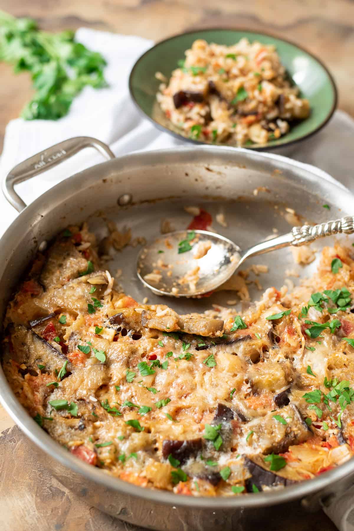 A serving spoon in a pan of baked eggplant and rice with a bowl of rice in the bacckground.