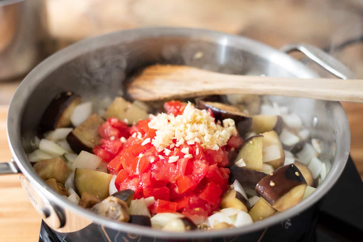 Chopped eggplant, tomatoes, and garlic with a wooden spoon in a pan on a cooktop.