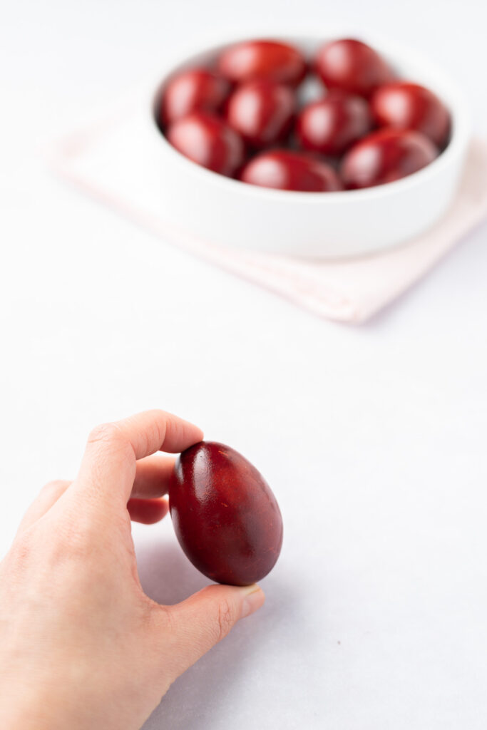 A hand holding a red-dyed egg with a bowl of red eggs in the background.